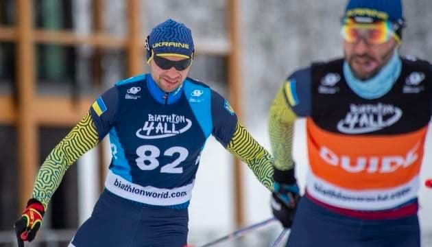 Two athletes compete in a biathlon cross-country skiing event. On the left, a skier wears a blue and yellow uniform with the number “82” and “UKRAINE” on the headband. The uniform also shows “BIATHLON 4ALL” and “biathlonworld.com.” On the right, a skier in a black, orange, and white vest labeled “GUIDE” accompanies the Ukrainian athlete, likely indicating visual impairment. The scene highlights inclusivity and teamwork in sports.