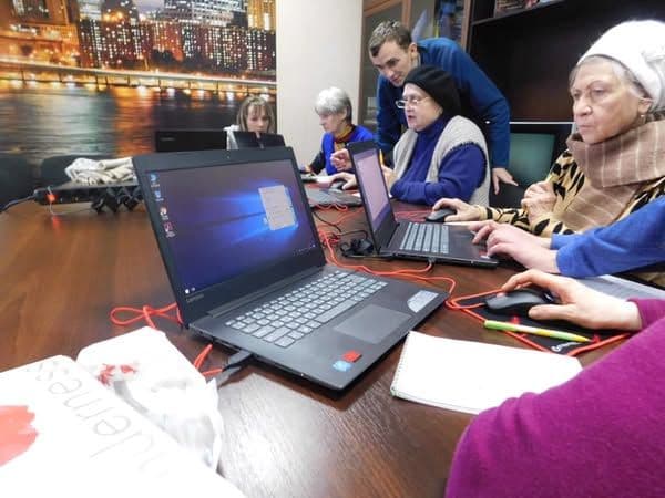 Elderly individuals are learning computer literacy, working on laptops. An instructor is assisting participants, explaining the tasks.
