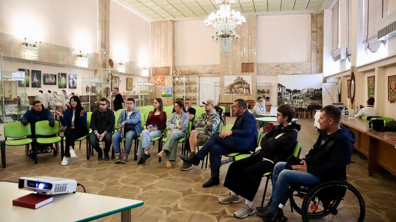 Дванадцять людей сидять півколом у просторій залі з високою стелею та люстрами, що нагадує музей або культурний центр. Уздовж стін — вітрини з експонатами та мистецькими роботами. Один із учасників — у візку, що підкреслює інклюзивність. На передньому плані — проектор на столі, спрямований на групу, що свідчить про презентацію або обговорення. Зелені стільці та невимушена атмосфера передають дух спільної участі та освітньої взаємодії.