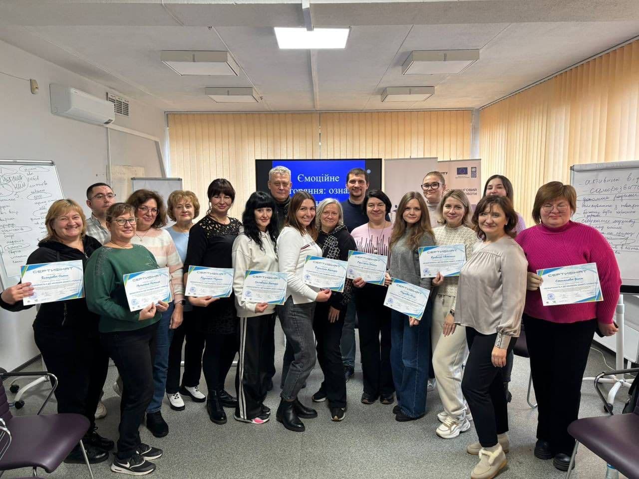 Group photo of training participants in a room with flip charts, a screen, and chairs. Most hold certificates. The screen displays “Emotional / Readiness: sign,” and the flip charts list terms related to emotional literacy and self-regulation. The scene captures the conclusion of an educational event focused on psychological readiness.