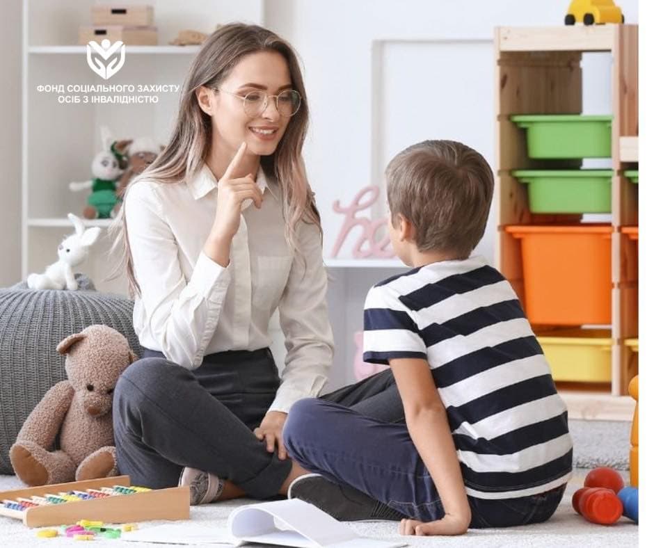 A woman and a child sit on the floor in a brightly lit room, resembling a classroom or therapy space. The woman, wearing a white blouse and glasses, engages with the child in a striped shirt. Around them are educational and play materials: a tray of colorful counting blocks, a teddy bear, and other toys. Behind them are shelves with colorful bins and stuffed animals, creating a child-friendly environment. The image includes the text “Social Protection Fund for Persons with Disabilities.” The scene conveys support and inclusivity in an educational or therapeutic setting.