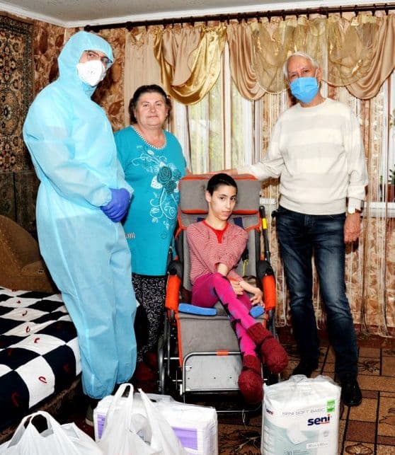 In a room with a window in the background, a girl sits in a specialized wheelchair, accompanied by two adults and a healthcare worker wearing protective gear, gloves, and a mask. In front of them are bags containing humanitarian aid, including hygiene products.