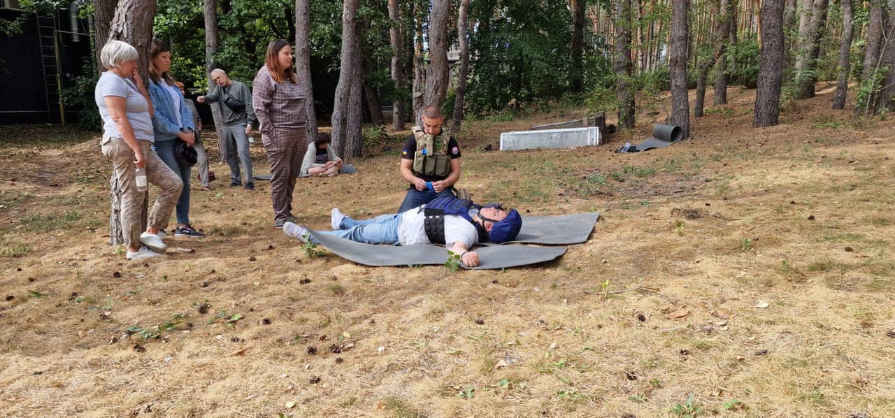 A man is lying on a mat, another man is crouching beside him helping, while three women are observing. In the background, a man is standing.