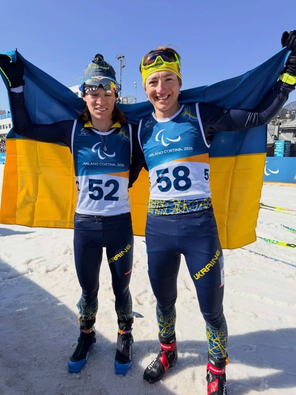 Two Ukrainian athletes stand on snow wearing sports uniforms with “UKRAINE” printed on their pants. They hold a large Ukrainian flag behind them. Their bibs display numbers “52” and “58” along with the Milano Cortina 2026 Winter Paralympics logo. Ski poles and a snowy sports venue are visible in the background. The scene conveys pride and achievement of Ukrainian Paralympians.