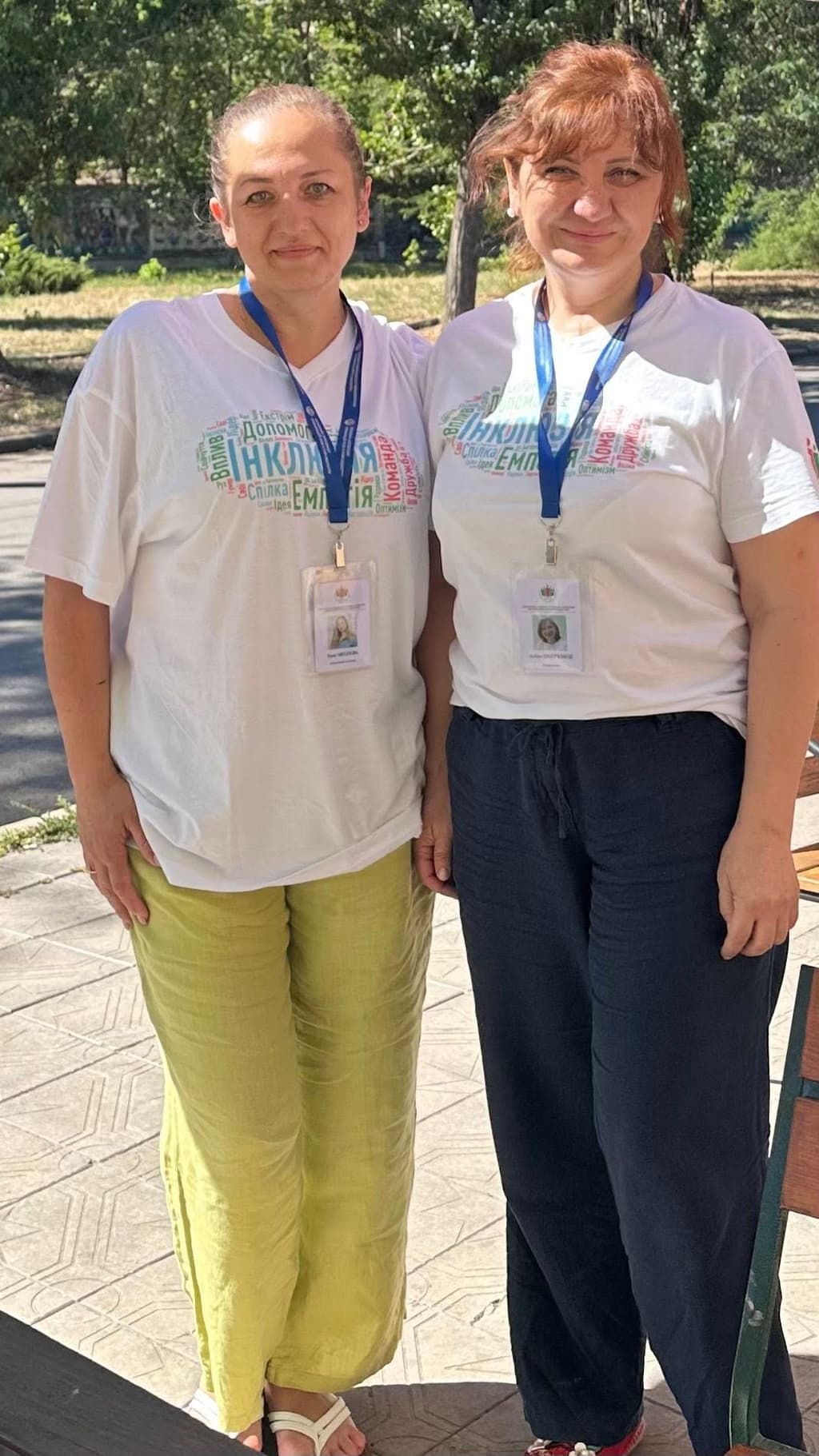 Two individuals stand outdoors side by side, both wearing white t-shirts with colorful Ukrainian text including “ІНКЛЮЗІЯ” and “ЕМІЛІЯ.” Each wears a blue lanyard with an ID badge. The person on the left has light green pants and white sandals; the one on the right wears dark blue pants and red shoes. They stand on a paved walkway flanked by trees, suggesting an event or gathering in a park-like setting that promotes inclusion.