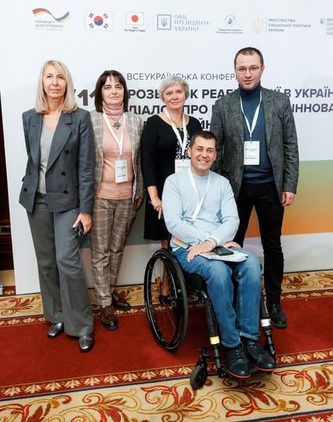 The image shows a group of five people posing for a photograph in front of a backdrop. The backdrop contains several logos and text in Ukrainian. One of the individuals is seated in a wheelchair, while the other four are standing. All individuals are wearing conference badges around their necks. The backdrop includes logos from various organizations, including the Ministry of Social Policy of Ukraine, the Office of the President of Ukraine, and other entities. The text on the backdrop is partially obscured but includes the phrase "ВСЕУКРАЇНСЬКА КОНФЕРЕНЦІЯ," which translates to "All-Ukrainian Conference."