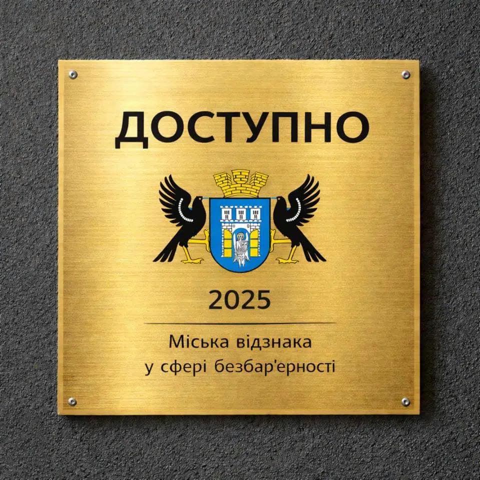 Золота табличка на темній фактурній стіні. У центрі — герб із двома чорними стилізованими птахами по боках. Над гербом напис «ДОСТУПНО», нижче — рік «2025». У нижній частині текст: «Міська відзнака у сфері безбар’єрності». Табличка символізує нагороду за досягнення у сфері доступності та інклюзивності.