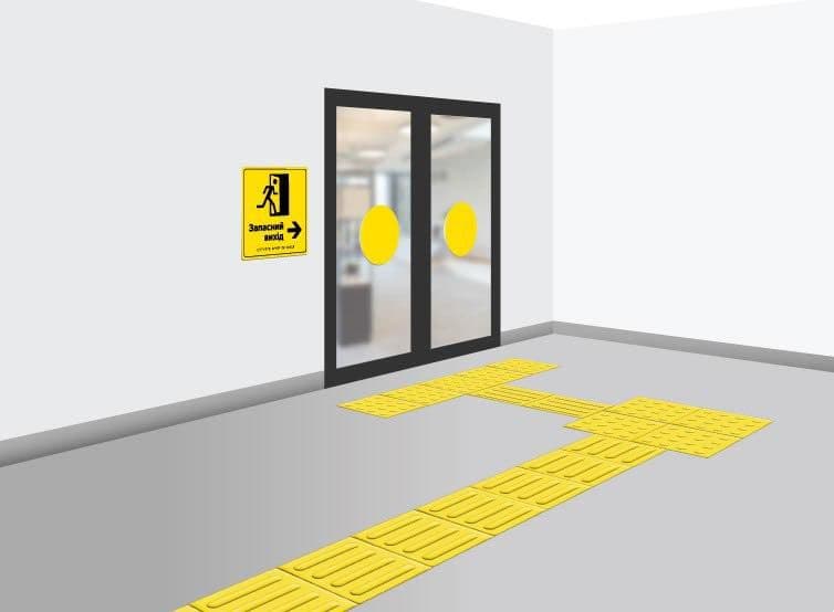 Коридор із яскравою жовтою тактильною плиткою, що веде до подвійних скляних дверей. Плитка має напрямні та Т-подібний перехрест, які допомагають людям із порушенням зору орієнтуватися. На стіні біля дверей висить жовта табличка з чорним текстом і піктограмою «Запасний вихід», зі стрілкою вправо. Сцена демонструє приклад доступності та аварійної навігації в громадській будівлі.