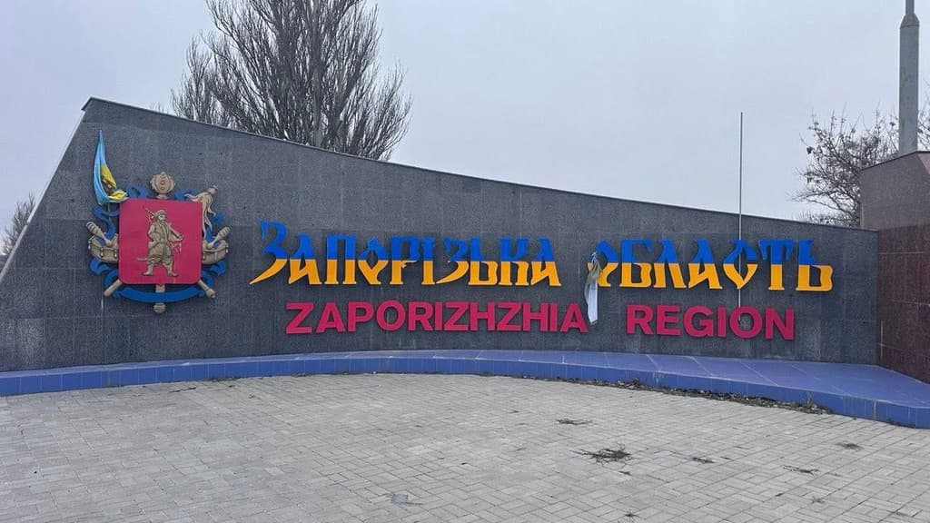 Великий придорожній знак на в’їзді до Запорізької області. Конструкція з темного каменю з кольоровими написами: «Запорізька область» синьо-жовтими літерами та «ZAPORIZHZHIA REGION» червоним. Ліворуч — герб із фігурою з зброєю, декоративними елементами та прапором України. На фоні — похмуре небо, голі дерева та асфальтована ділянка. Сцена позначає адміністративну межу регіону.