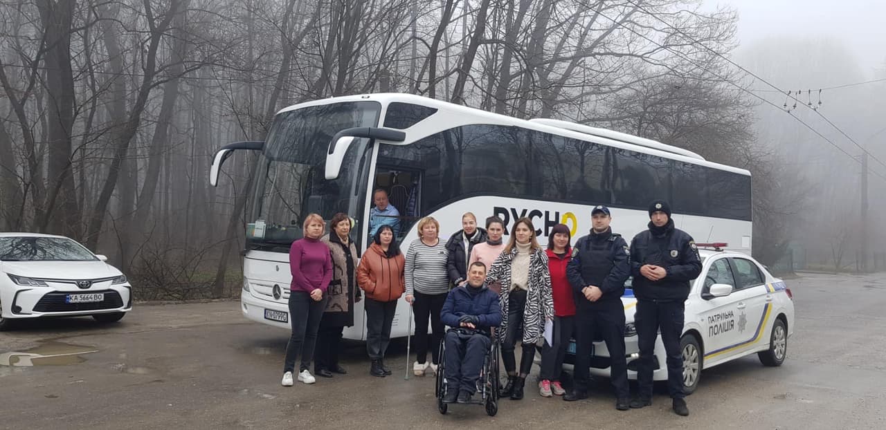 На зображенні група людей стоїть перед великим автобусом з написом "РУСЧО". Поруч з автобусом стоїть поліцейський автомобіль з написом "ПАТРУЛЬНА ПОЛІЦІЯ". Деякі люди одягнені в зимовий одяг, а один з них сидить у інвалідному візку. На задньому плані видно дерева в тумані.