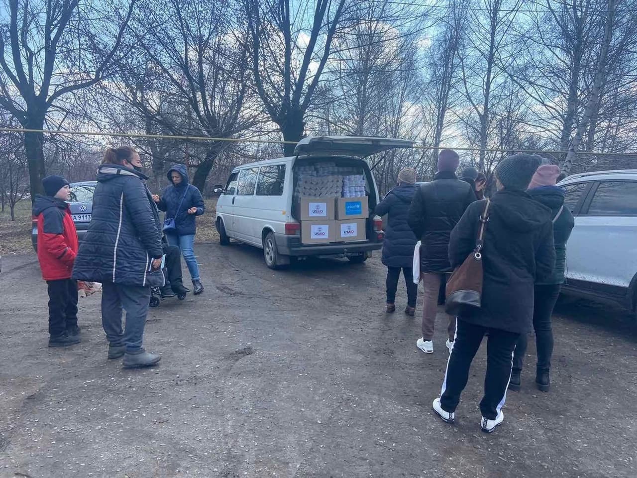 Група людей зібралася біля білого фургона з відчиненими задніми дверима. Усередині видно коробки та пакунки з логотипами «USAID» і «UNICEF». Люди в зимовому одязі стоять поруч, деякі чекають у черзі чи отримують допомогу. На задньому плані — голі дерева та припарковані автомобілі, що свідчить про холодну пору року та сільську чи приміську місцевість. Сцена демонструє процес розподілу гуманітарної допомоги міжнародними організаціями.