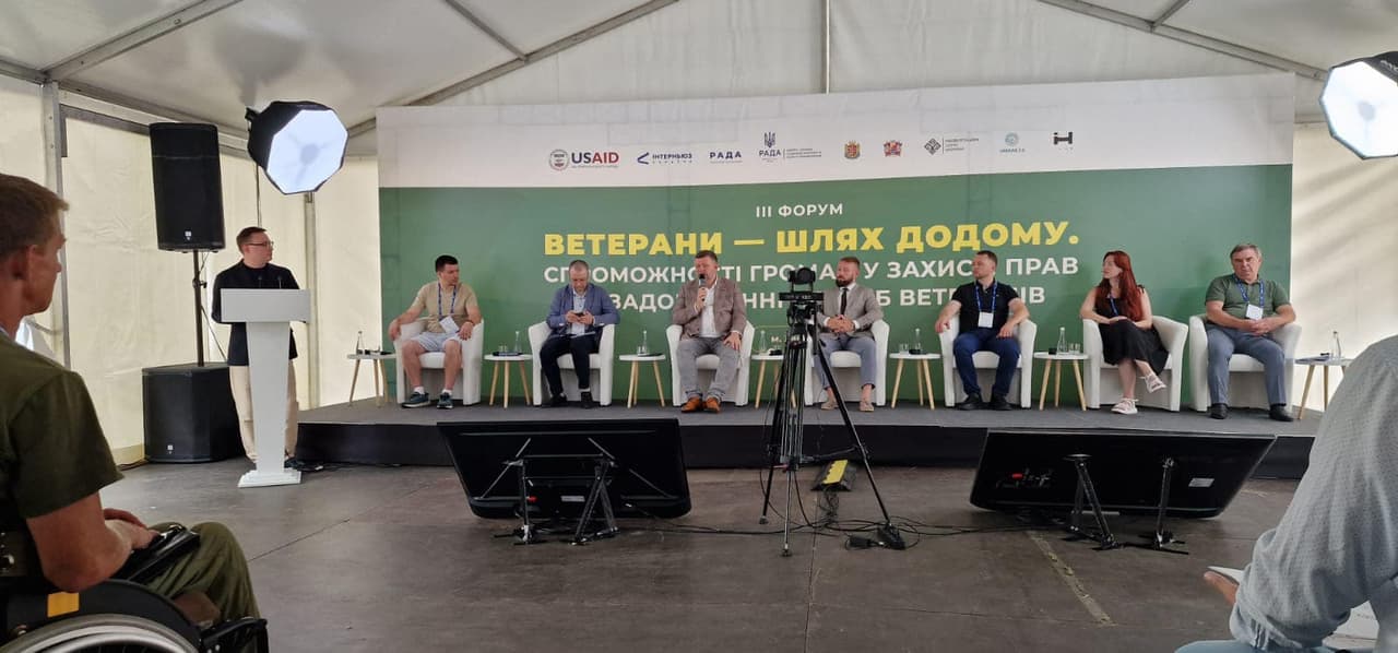 Сім людей сидять на фоні банера на платформі, один чоловік доповідає за стійкою, по краям зображено глядачів.