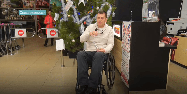A man in a wheelchair is seated near an information desk with a Christmas tree in the background inside a store. A woman is walking behind him. The top-left corner displays the text "Sievierodonetsk.