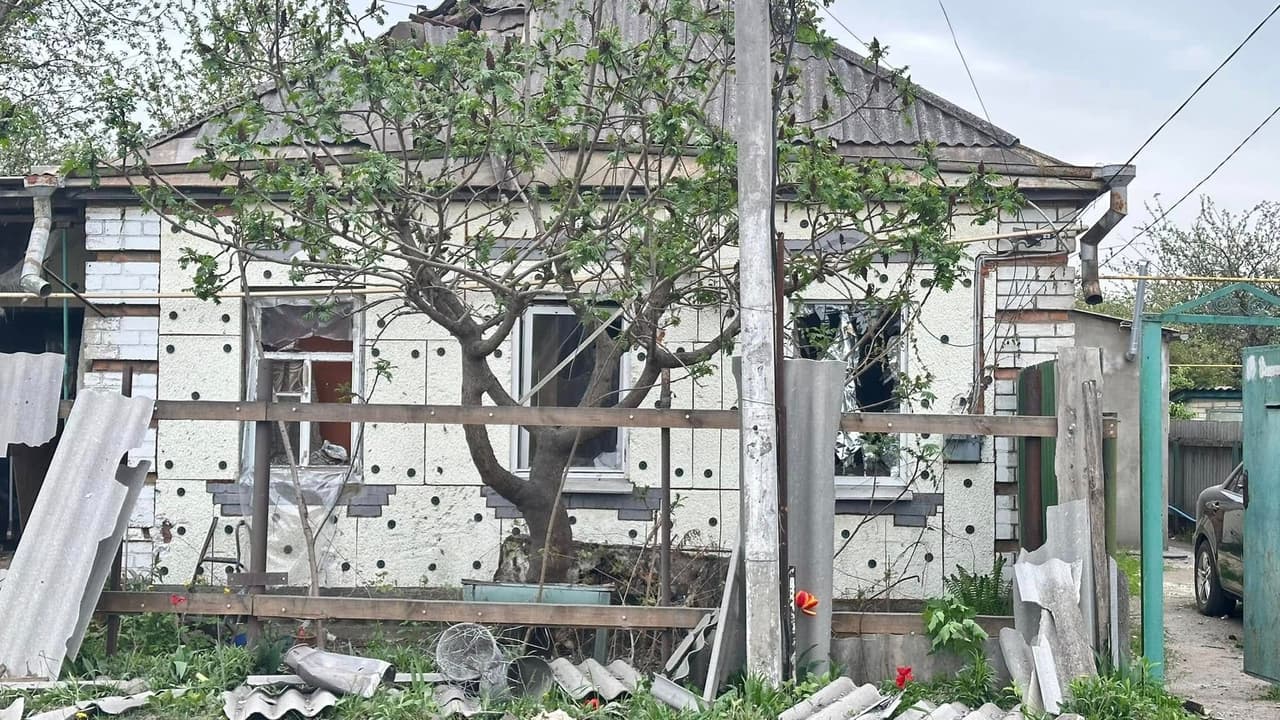 На зображенні зображений будинок із видимими пошкодженнями та ознаками занедбаності. До будинку дуже близько росте дерево, гілки якого закривають вікна. Зовнішні стіни частково вкриті ізоляційним матеріалом із чорними круглими плямами, а деякі частини стіни залишаються відкритими, де видно цегляну кладку. У будинку розбиті вікна, а на передньому дворі розкидані різні будівельні матеріали та сміття, включаючи металеві листи та дерев'яні дошки. Перед будинком знаходиться бетонний стовп, а з правого боку видно частину автомобіля. Сцена натякає на те, що будинок може перебувати у стані ремонту чи пошкодження.