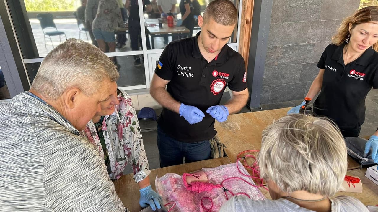 A girl and a boy, instructors in gloves, along with three other people, stand by a table with training models for providing assistance in case of injuries.