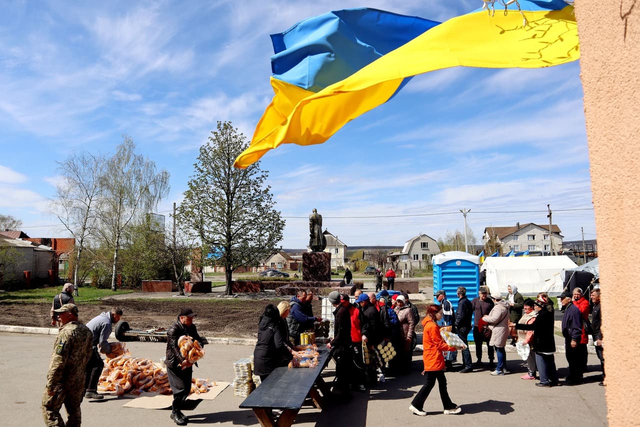 Люди стоять у черзі за гуманітарною допомогою на тлі українського прапора, що майорить на вітрі. Роздача хліба та інших продуктів у прифронтовій зоні.