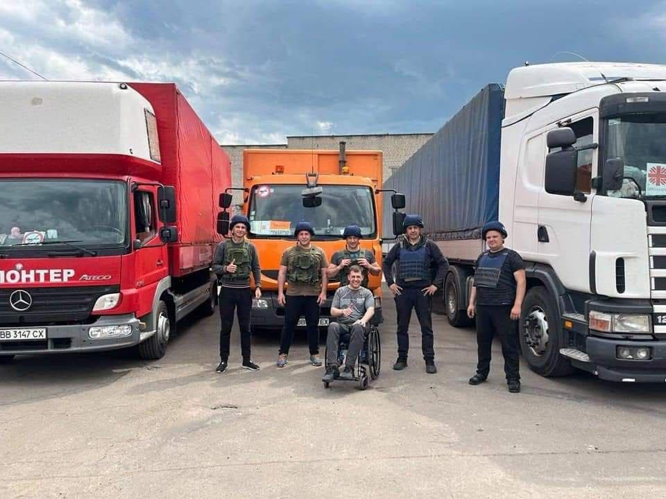 Six individuals stand in front of three large trucks—red, orange, and white. All wear military or tactical gear, including helmets and body armor. A man in a wheelchair is positioned at the center. The red truck displays the word “ЮНТЕР” and license plate “BB 3147 CX.” The white truck features a logo resembling a humanitarian organization. A brick building and cloudy sky form the backdrop. The scene suggests coordination or support, possibly in a humanitarian or logistics context.