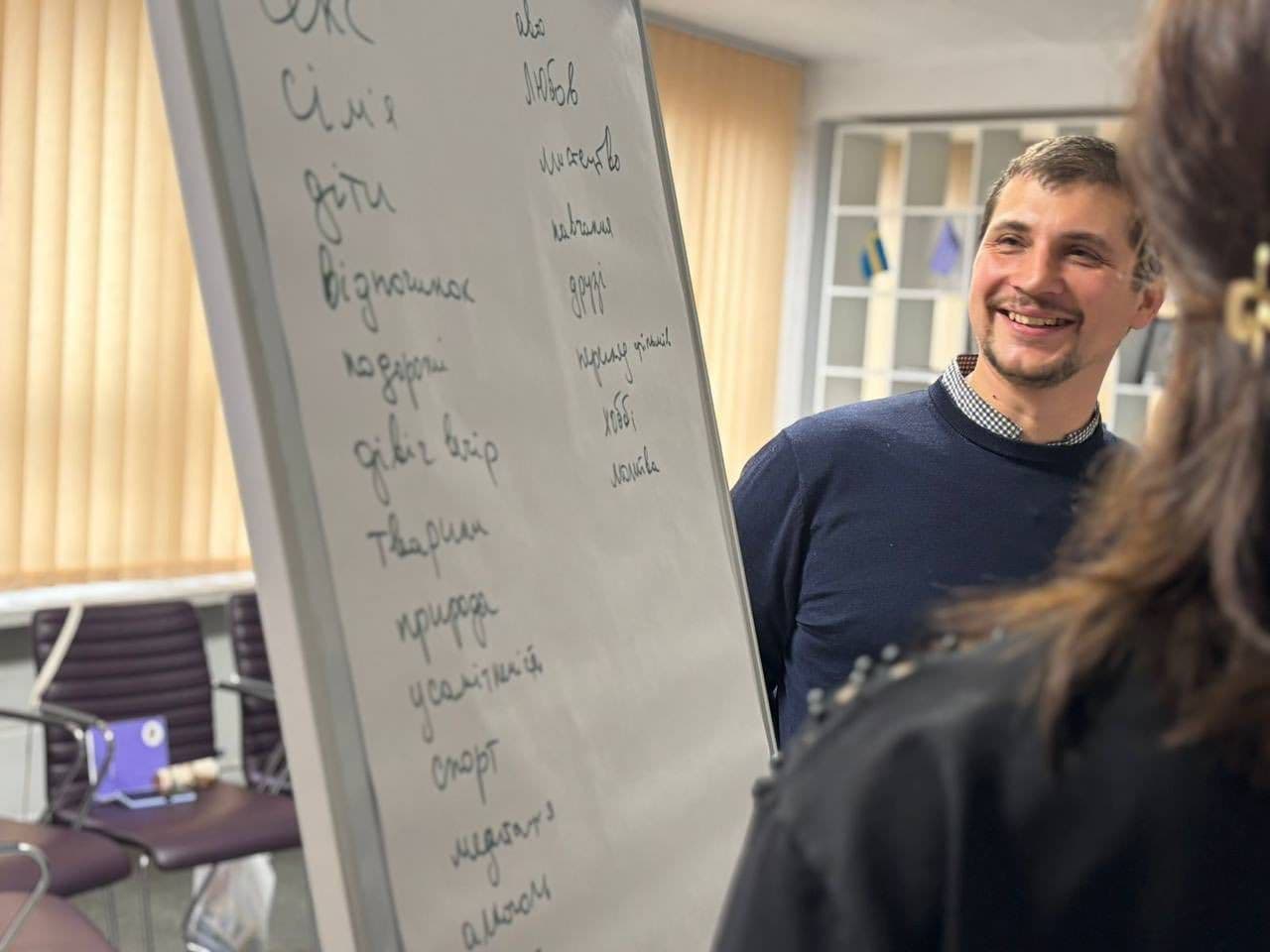 A man stands next to a whiteboard with handwritten Ukrainian words, speaking to another person seated with their back to the camera. The room has vertical blinds and chairs arranged in a semi-circle. The board lists words like “family,” “children,” “rest,” “love,” “art,” and “money.” The scene suggests a workshop or group discussion on values or sources of personal resilience.