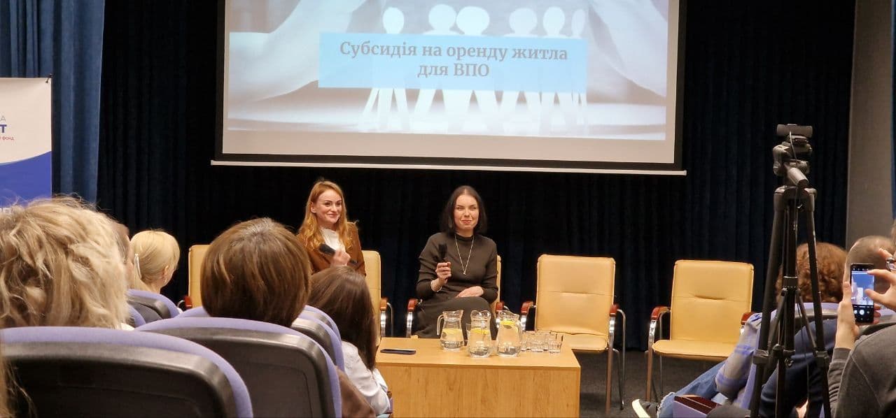 Presentation on Housing Subsidies for IDPs: Two women sit on stage in a conference hall, holding microphones, during a presentation on “Housing Subsidy for IDPs” (internally displaced persons). The title of the presentation is displayed on a blue background on the projector screen. The audience is seated in the hall, with some recording the event on their phones.