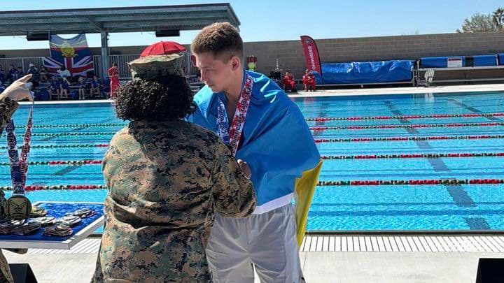 Церемонія нагородження на змаганнях із плавання. Військовослужбовець у камуфляжній формі вручає медаль спортсмену, загорнутому в синьо-жовтий прапор України. На шиї плавця вже кілька медалей. Вони стоять біля басейну з доріжками. На задньому плані — глядачі, прапори (зокрема британський Union Jack) та банери змагань, що свідчить про міжнародний рівень події. Сцена передає момент досягнення та визнання.