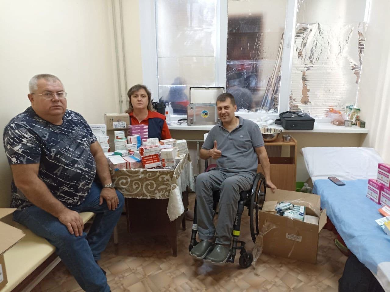 Three people are in a small room resembling a medical or aid distribution space. One man in a wheelchair gives a thumbs-up gesture, while two others sit nearby. A table with a patterned cloth is covered with medical supplies and pharmaceutical boxes. A cardboard box with more items sits on the floor, and additional supplies are stacked on a bed or examination table. The window is covered with reflective material, and various items are placed on the windowsill and a cabinet. The scene reflects a moment of humanitarian or medical support in a temporary or field setting.