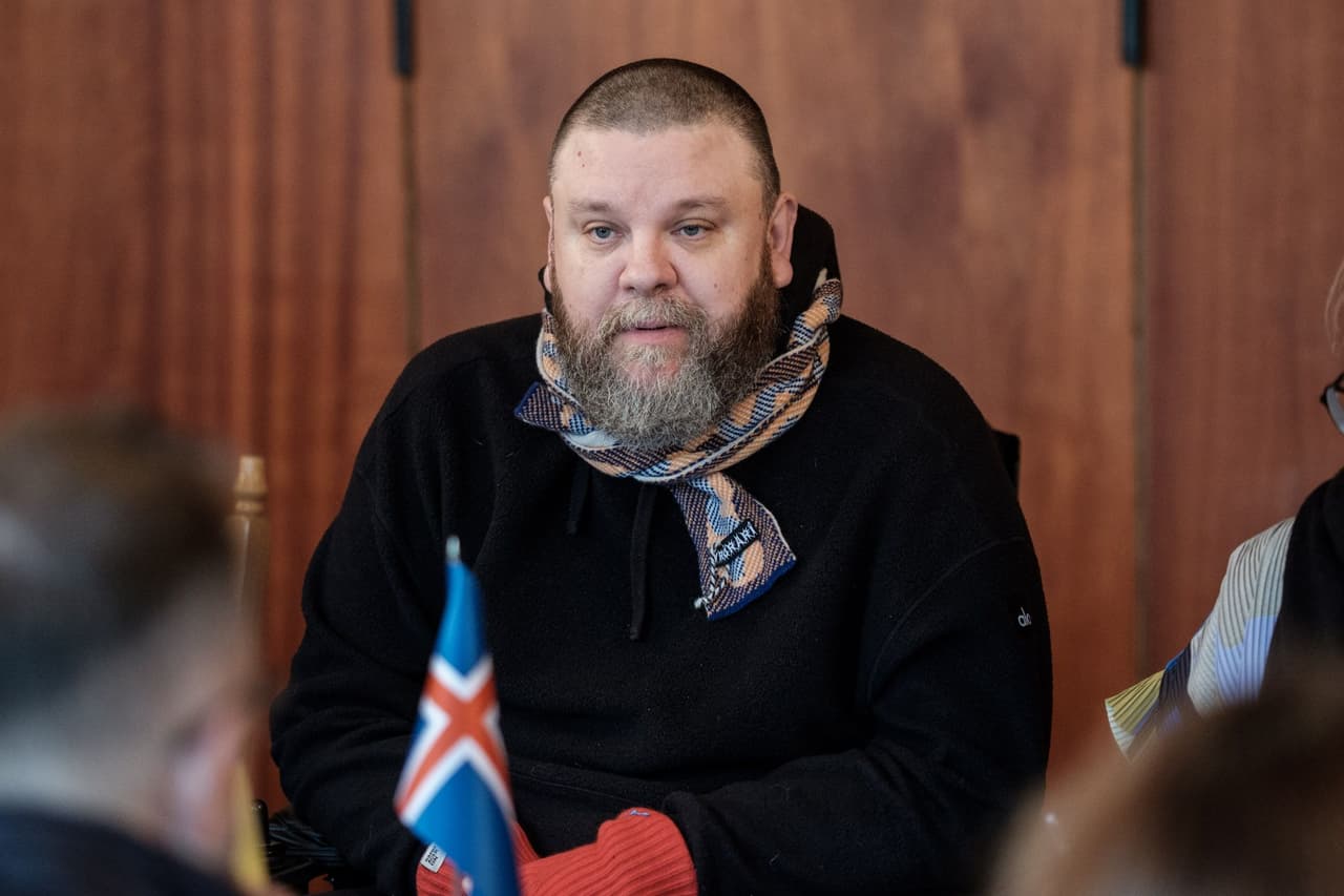 Людина з бородою, вдягнена в чорний светр, різнокольоровий шарф і червоні рукавички, сидить у приміщенні з дерев’яними панелями на стіні. Перед нею — маленький прапор Ісландії, що може свідчити про національну приналежність або контекст події. Поруч частково видно інших учасників, що створює атмосферу формальної або напівофіційної зустрічі. Сцена передає міжнародну присутність і інклюзивність у професійному середовищі.