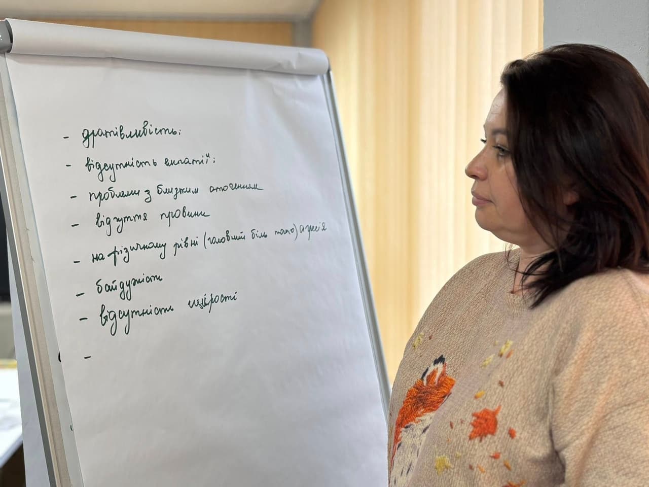 A woman stands next to a flip chart with handwritten Ukrainian text, likely leading a presentation or workshop. The chart lists signs of emotional exhaustion, including irritability, lack of empathy, strained relationships, guilt, physical symptoms (e.g., headaches), aggression, indifference, and lack of sensitivity. The setting appears to be educational or training-oriented.