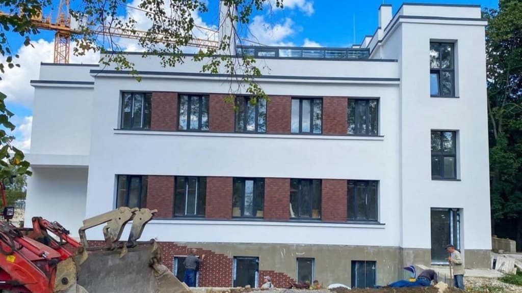 На зображенні сучасна багатоповерхова будівля на етапі будівництва. Фасад білий із секціями червоної цегли навколо вікон, що створює контрастний дизайн. Видно великі прямокутні вікна, розташовані у сітчастому порядку на трьох поверхах. У нижній частині будівлі працюють будівельники, які встановлюють цеглу. На передньому плані стоїть червоний екскаватор із великою ковшовою лопатою. На задньому фоні видно будівельний кран, дерева та блакитне небо з хмарами. Атмосфера передає процес активного будівництва.