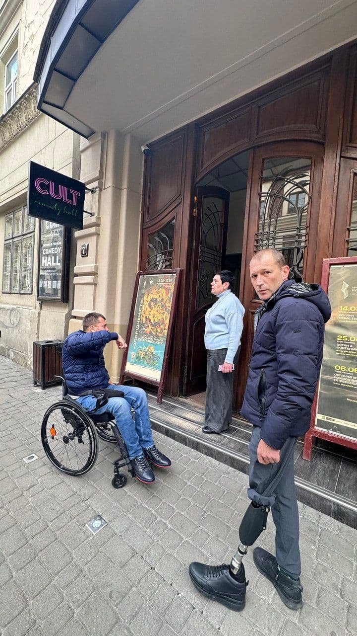 The entrance of a building labeled "CULT comedy hall." Three people are present: one in a wheelchair, one standing with a prosthetic leg, and another near the doorway. Two posters are displayed at the entrance—one featuring a colorful scene with foreign language text and another listing dates and events. The setting emphasizes accessibility and inclusion in entertainment spaces.