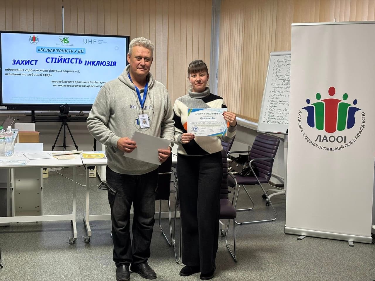 A man and woman stand in a training room. The woman holds a certificate stating “Kurashova Lina successfully completed the program ‘Barrier-Free in Action: Protection, Resilience, Inclusion.’” Behind them is a presentation slide with the program title and logos of LAOOI, UHF, and partner organizations. A flip chart with notes and a banner of the Luhansk Association of Organizations of Persons with Disabilities are also visible. The scene marks the completion of a training for professionals in social, educational, and medical fields.