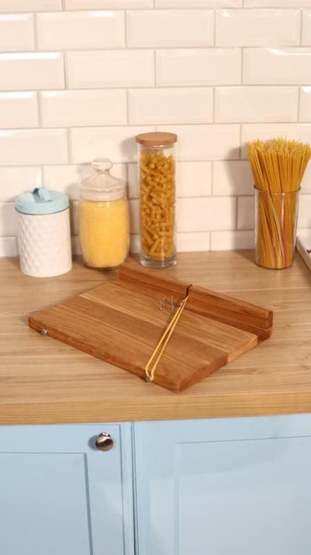 На кухонній стільниці стоїть дерев’яний пристрій, схожий на саморобний інструмент для вимірювання порцій спагеті. Він складається з плоскої основи, піднятої секції та металевих гачків, між якими натягнута жовта гумка. Позаду розташовані чотири ємності: біла керамічна банка з синьою кришкою, скляна банка з жовтою крупою (схожою на кукурудзяну), висока банка з макаронами «локшина-ріжки» та ще одна висока банка зі спагеті. На фоні видно білу плитку «кабанчик» та світло-блакитні шафки під стільницею.