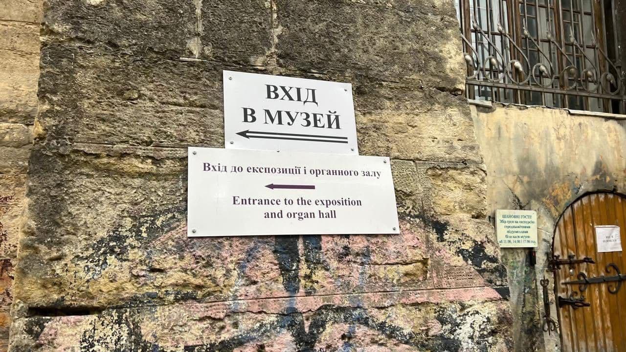 A stone wall with multiple directional signs pointing to a museum, exposition, and organ hall. The signs are in both Ukrainian and English. Graffiti is visible at the bottom, and to the right is a wooden door with iron bars and hinges, topped by a barred window. The scene captures a historical or cultural site with bilingual signage for visitors.