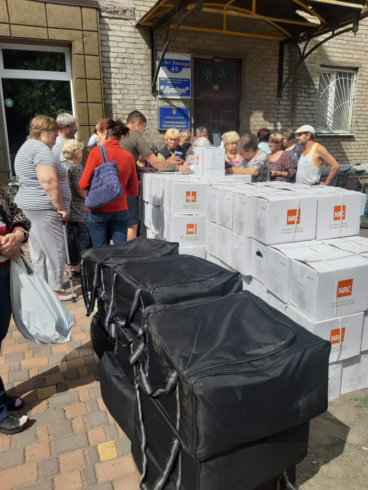People stand in line in front of the building of the public organization “Slovyansk City Society of Disabled People” to receive humanitarian aid. In the foreground, there are black thermal bags and stacks of white boxes with the NRC (Norwegian Refugee Council) logo. Volunteers are registering and distributing aid.