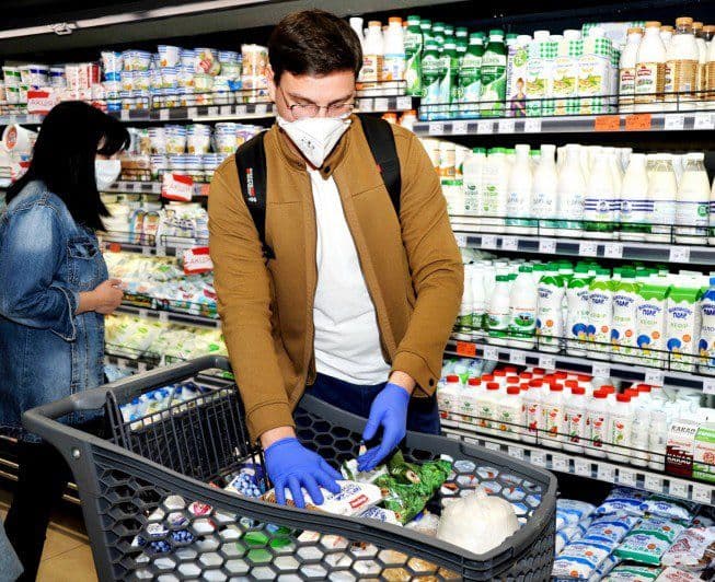 A man wearing a protective mask and gloves is shopping in a supermarket. He is placing groceries, including vegetables and dairy products, into a large shopping cart. In the background, shelves filled with dairy products are visible, and a woman in a denim jacket is also shopping nearby.