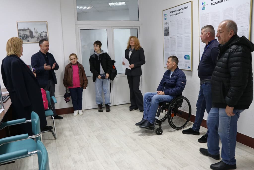 Eight people gather in a bright room with white walls and light flooring, resembling a public service or community center. Teal chairs line the left wall, and large posters with Ukrainian text are displayed on the right, including one titled “TAXES AND LAW.” One person is seated in a wheelchair, while others stand and engage in discussion. A woman holding papers appears to be presenting or reading aloud. The setting suggests an informational session or community meeting focused on legal or civic topics, with an inclusive atmosphere.