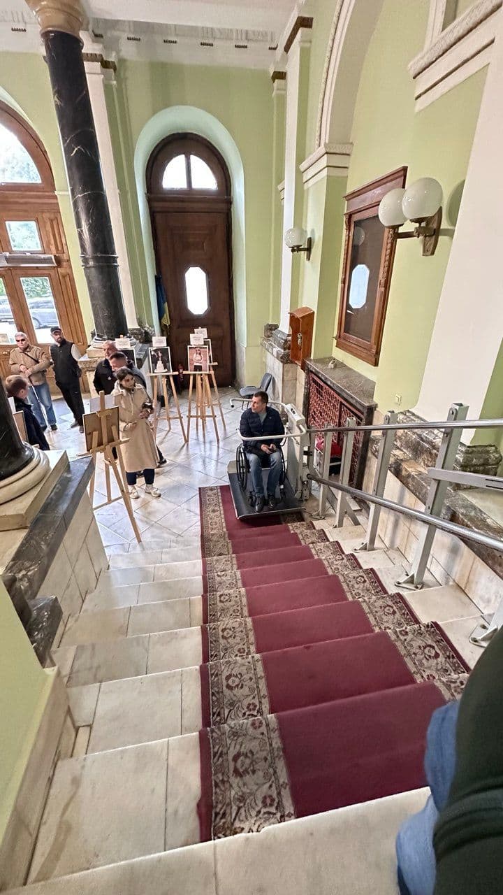 The interior of a building with a grand staircase covered by a red carpet featuring intricate patterns. A person in a wheelchair is using a stairlift to descend the stairs, which is attached to the right-hand railing. Several people are present, some engaged with easels displaying photographs or artwork. The architecture includes high ceilings, large arched windows, and a wooden door with a glass panel. The walls are painted light green, with marble columns and decorative details, suggesting a historical or public building, possibly hosting an exhibition or event.