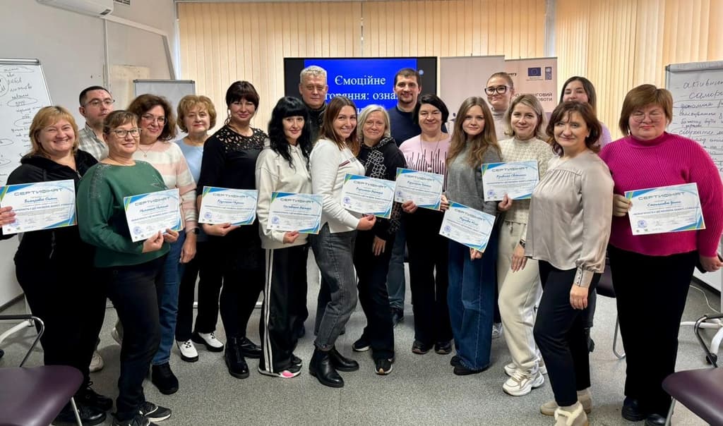 Групове фото учасників та учасниць тренінгу з психосоціальної підтримки. Люди стоять у два ряди в навчальній аудиторії та тримають у руках сертифікати про проходження навчання. На задньому плані — фліпчарти та екран із темою заняття.»