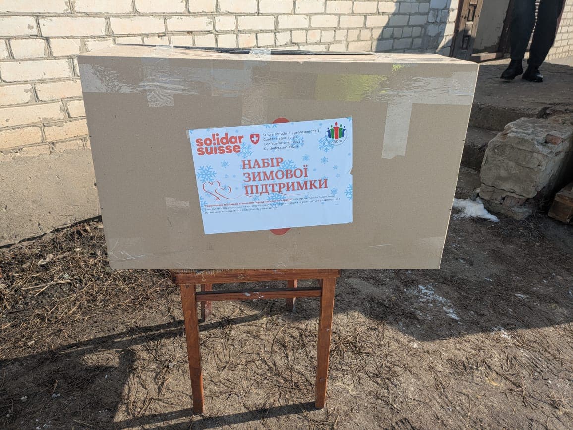 A large box labeled “Winter Support Kit” sits on a table near a building. It is part of humanitarian aid for people in difficult conditions.