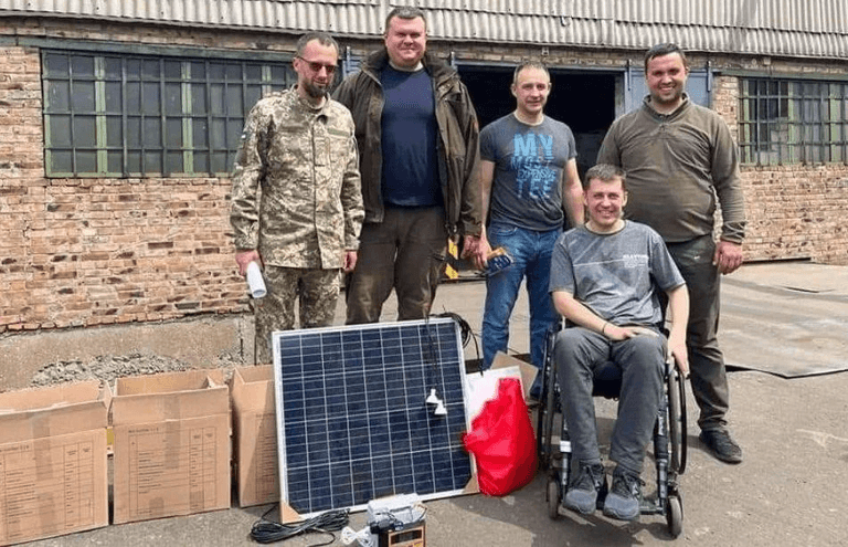 Five men near a solar panel against the backdrop of a one-story brick building