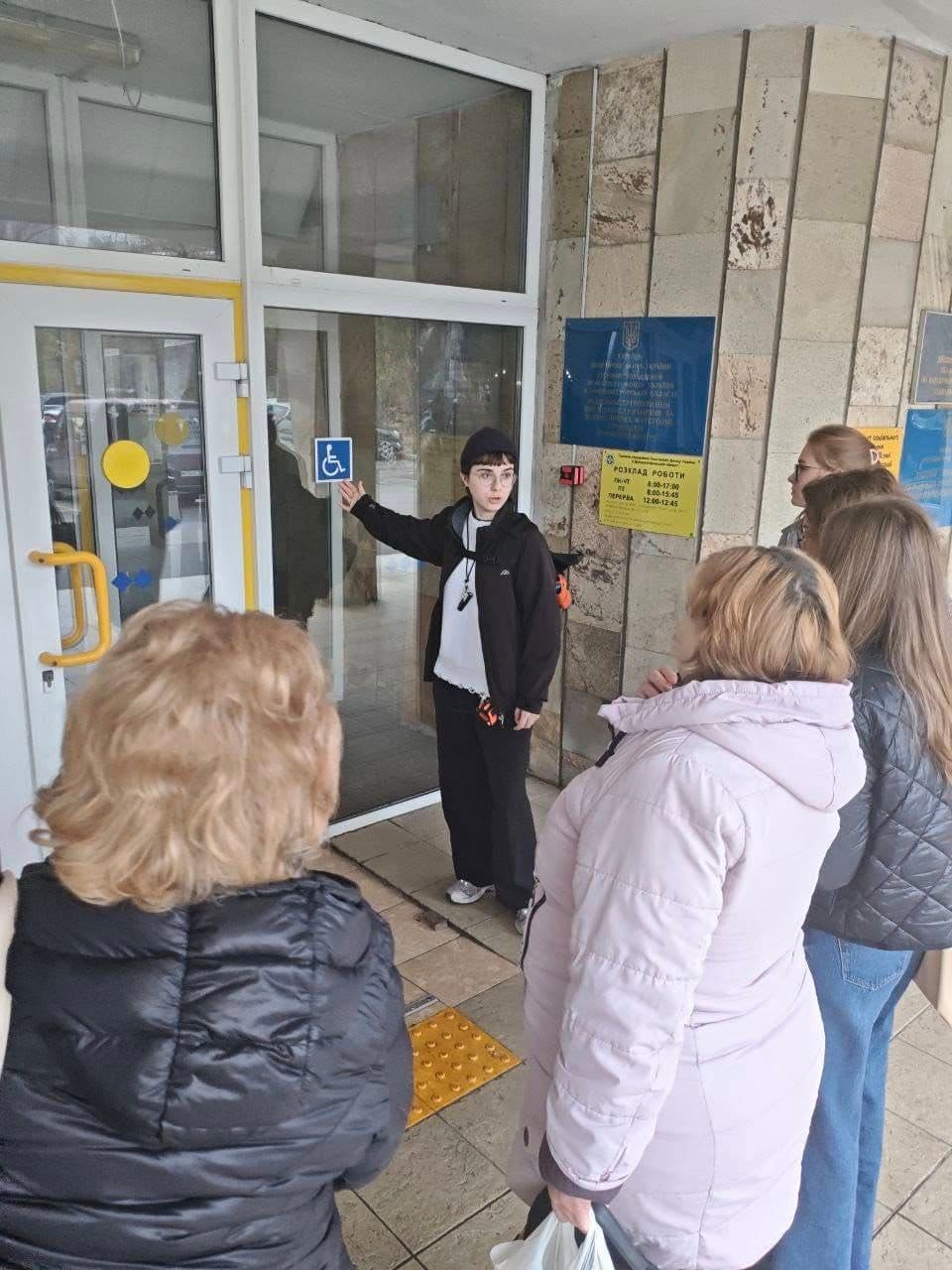 A group of people stands near a building entrance. One person points to a wheelchair symbol indicating accessibility. The glass doors have yellow handles, and tactile paving is visible on the ground. Informational signs are posted on the wall, including a Ukrainian-language schedule. The scene highlights features of an accessible public space.