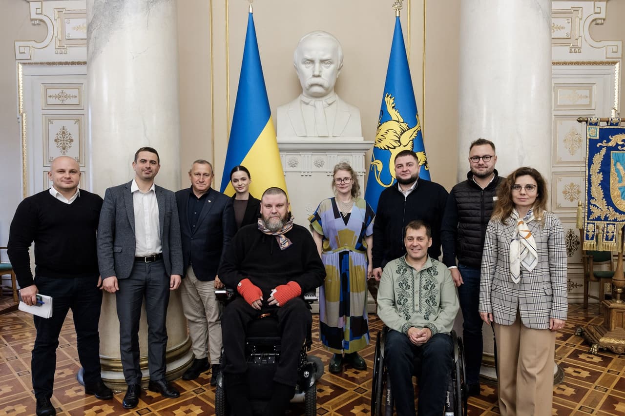 Одинадцять людей позують у пишному приміщенні з класичною архітектурою — білими колонами та декоративними елементами на стінах. Попереду — двоє осіб у візках, позаду — решта учасників стоять. За групою — білий бюст чоловіка на стіні, між державним прапором України та синім прапором із золотим тризубом. Обстановка передає урочисту атмосферу, ймовірно, пов’язану з державною діяльністю, адвокацією або вшануванням, з акцентом на інклюзивність і громадську участь.