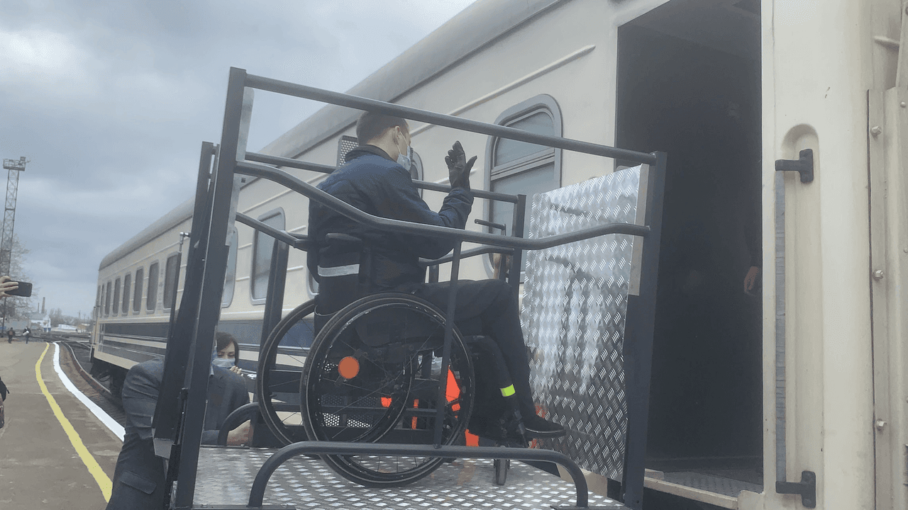 Людина у візку піднімається на потяг за допомогою металевого платформеного підйомника. Потяг стоїть на платформі, а поруч знаходяться інші люди, які допомагають або спостерігають за процесом. Підйомник для візків розроблений, щоб допомогти людям із порушенням мобільності безпечно та комфортно сісти на потяг. Це зображення підкреслює важливість доступності у громадському транспорті, забезпечуючи інклюзивність для осіб з інвалідністю.