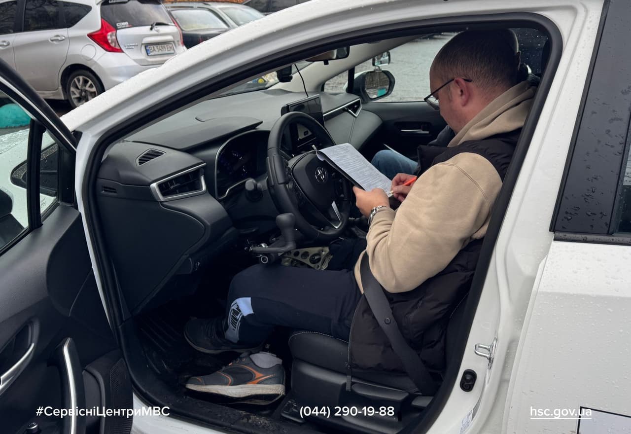 Людина в білому автомобілі заповнює документи, тримаючи аркуш і червону ручку. Водій одягнений у бежеву худі та чорну жилетку. У салоні — сучасна панель керування, кермо та важіль перемикання передач. На пасажирському сидінні — ще одна особа. Авто припарковане на стоянці, де видно інші машини. Сцена нагадує адміністративну процедуру, ймовірно, пов’язану з реєстрацією або оглядом транспортного засобу.