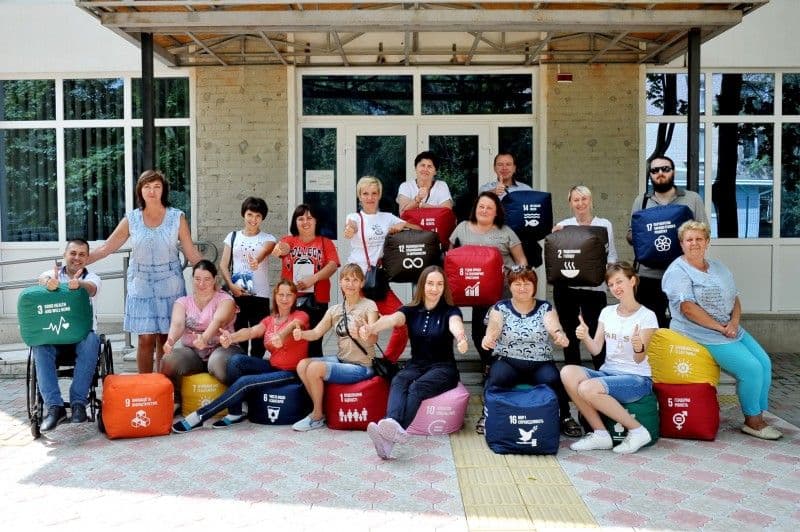 A group of people of different ages and genders sits in front of a building with glass doors and windows. They are holding colorful cubes representing the UN Sustainable Development Goals (SDGs). In the foreground, people smile and give thumbs up. In the background, a few more people are also holding cubes. A wheelchair-accessible ramp is visible.