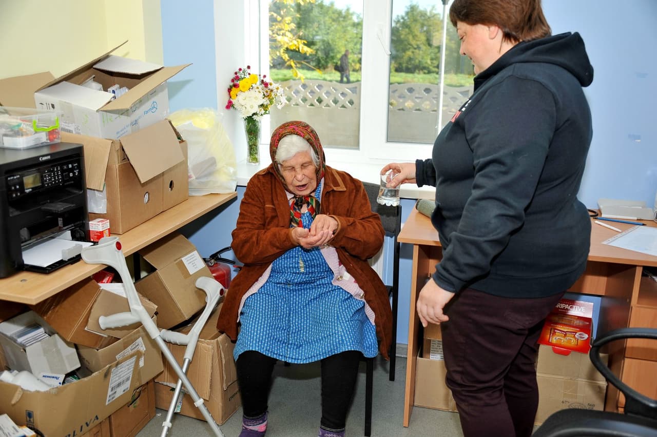 The image shows two people in a room that appears to be cluttered with various items. One person is seated and appears to be elderly, wearing a headscarf, a blue dress, and a brown jacket. The seated person is holding something in both hands. The other person is standing and holding a glass of water, seemingly offering it to the seated person. The room contains several cardboard boxes, some of which are open and filled with various items, including medical supplies. There is a printer on a desk, and a pair of crutches leaning against the desk. A vase with flowers is placed on the windowsill, and there is a window in the background showing an outdoor scene with greenery and a person walking in the distance.