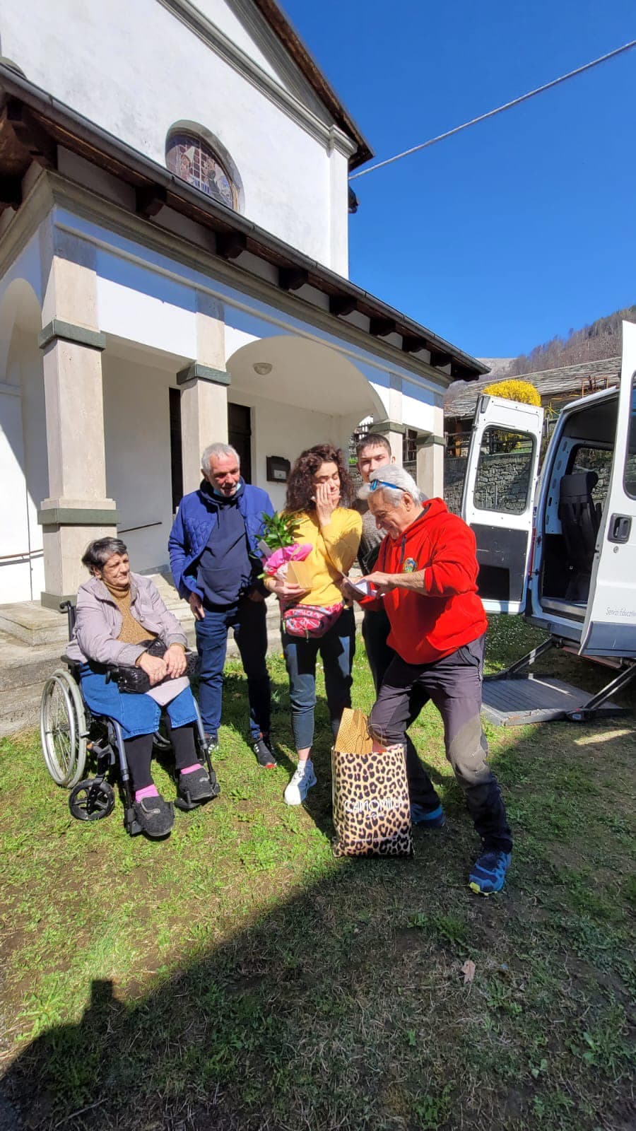 На зображенні видно групу з чотирьох людей, які стоять біля будівлі з білими стінами та арковими вікнами. Один із них сидить у інвалідному візку, а троє стоять поруч. Особа в червоній куртці передає щось особі в жовтій сорочці, яка тримає горшкову рослину, загорнуту в рожевий папір. На задньому плані видно білий фургон із відкритими дверима, що вказує на те, що група може брати участь у якійсь зовнішній діяльності чи події. Сцена відбувається на трав'янистій території під ясним блакитним небом.