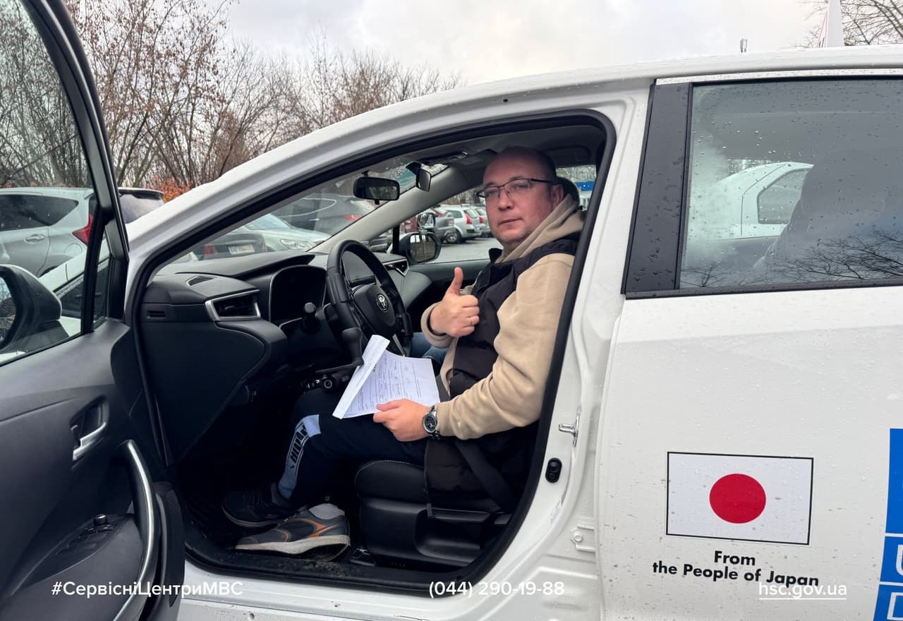 Чоловік сидить за кермом білого автомобіля з відкритими дверима, тримає документи та показує жест «палець вгору». На дверцятах — наліпка з прапором Японії та написом «From the People of Japan». На задньому плані — автостоянка з іншими машинами та голими деревами, що свідчить про холодну пору року. Сцена нагадує адміністративну процедуру, пов’язану з транспортом.
