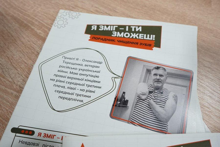 На зображенні буклет лежить на дерев’яній поверхні. На обкладинці — фотографія чоловіка без рук, у смугастій майці. Текст українською мовою містить мотиваційне повідомлення: «Я ЗМІГ – І ТИ ЗМОЖЕШ! ПОРАДНИК. ЧИЩЕННЯ ЗУБІВ». У «хмаринці» з промовою він представляється: «Привіт! Я – Олександр Терещенко, ветеран російсько-української війни. Маю ампутацію правої верхньої кінцівки на рівні середньої третини плеча, лівої – на рівні середньої третини передпліччя». Нижня частина буклету частково закрита, але видно повторення фрази «Я ЗМІГ – …».