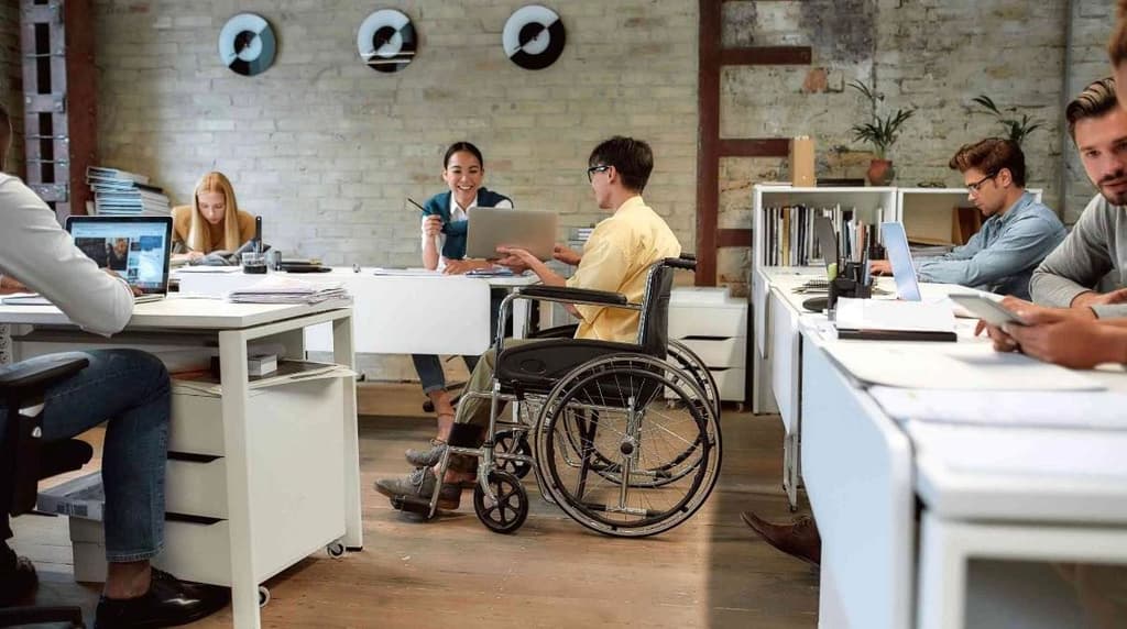 A modern office environment with several people working at white desks using laptops and tablets. One employee in a wheelchair converses with a smiling colleague holding a pen. Other workers are focused on their tasks. The background features a brick wall with decorative elements, shelves with books, and plants. The scene highlights inclusivity, diversity, and accessibility in a professional workplace.