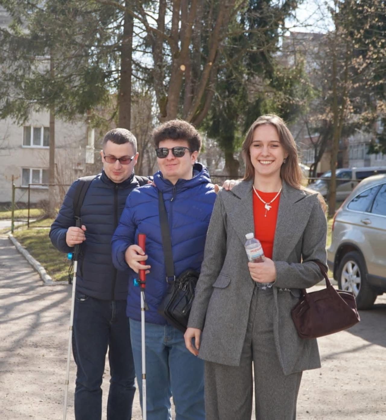 На зображенні троє людей стоять на вулиці сонячного дня. Двоє з них, ліворуч, тримають білі тростини, що свідчить про порушення зору. Вони одягнені у сині куртки та носять сонцезахисні окуляри. Праворуч стоїть жінка у сірому костюмі з червоною блузою, тримає пляшку води «Моршинська» та бордову сумку. На задньому фоні видно дерева, припаркований автомобіль та житлові будинки, що створює атмосферу міського середовища.